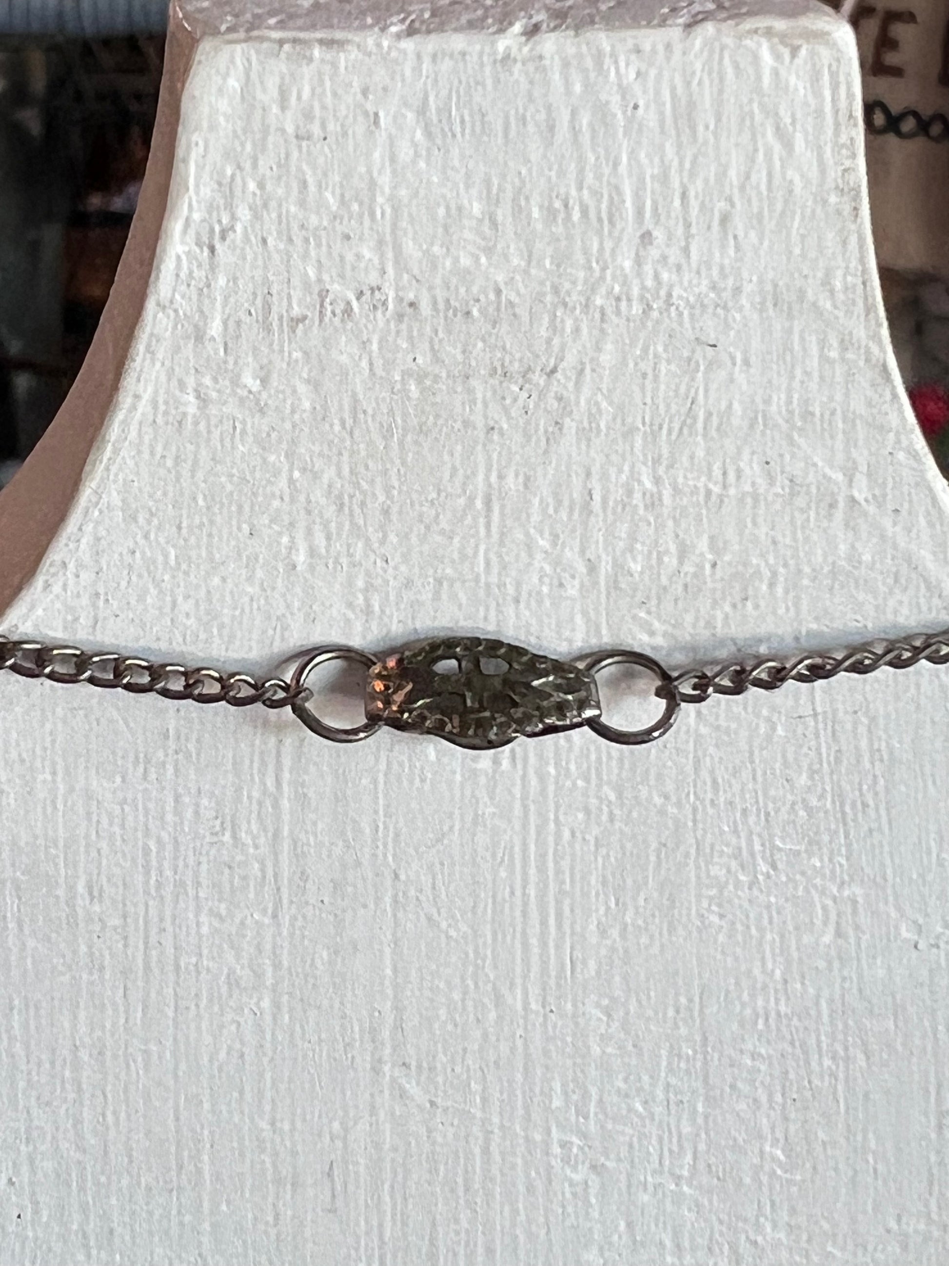 A silver chain bracelet with a textured, organic-shaped centerpiece and two small rings, displayed on a white textured bust.