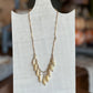 A gold-toned necklace with multiple cream-colored teardrop beads, displayed on a white fabric mannequin bust against a blurred indoor background.