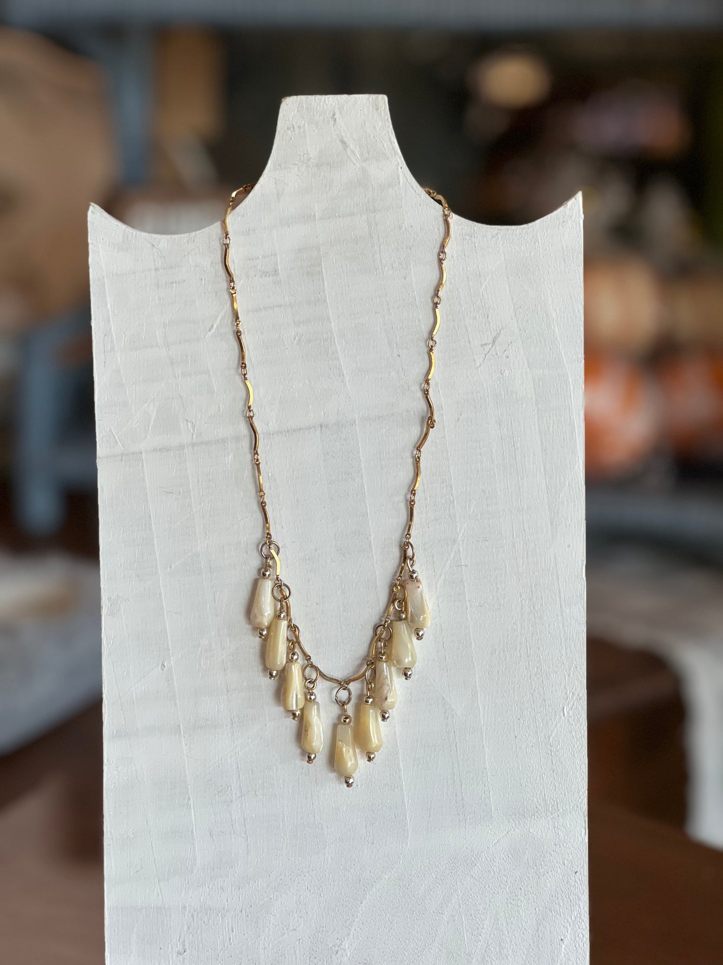 A gold-toned necklace with multiple cream-colored teardrop beads, displayed on a white fabric mannequin bust against a blurred indoor background.