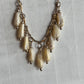 A vintage-style statement necklace featuring multiple cream-colored teardrop beads with a gold-toned chain and small metallic accents, laid flat on a textured white fabric background.