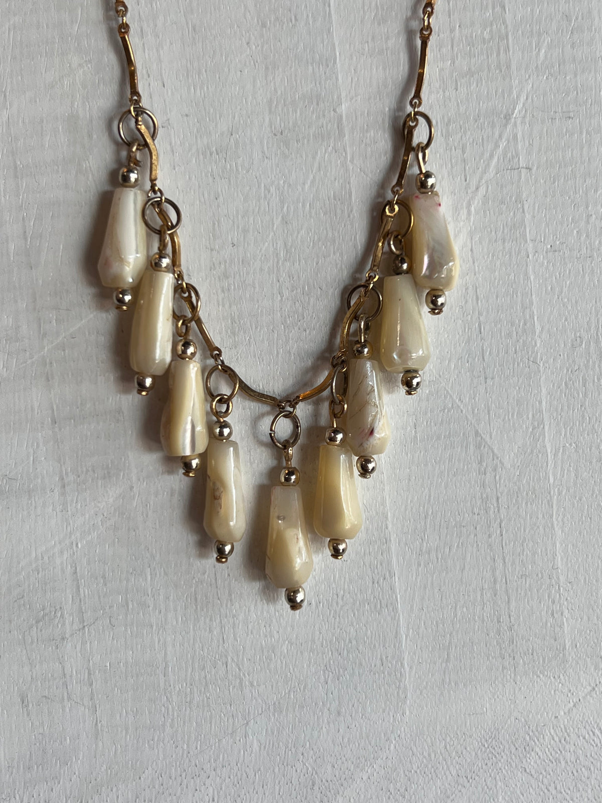 A vintage-style statement necklace featuring multiple cream-colored teardrop beads with a gold-toned chain and small metallic accents, laid flat on a textured white fabric background.