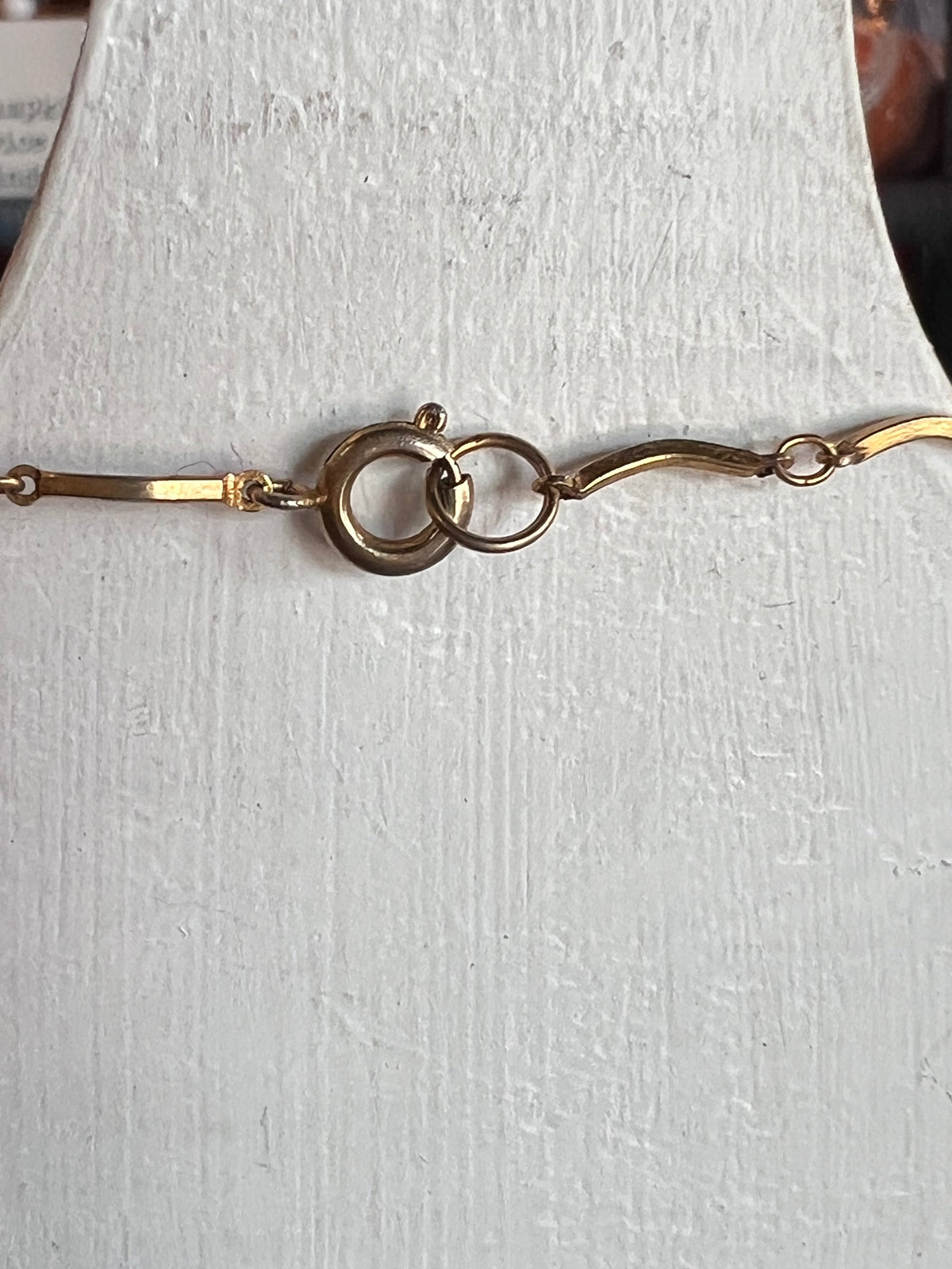A close-up of a gold-toned metal chain bracelet or necklace with a lobster clasp and interlocking ring detail, displayed on a textured white surface.