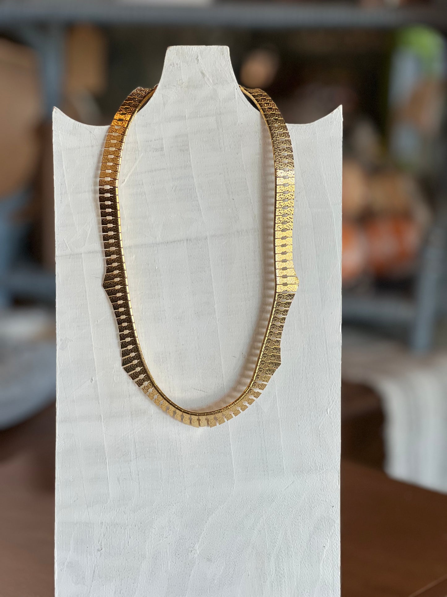 A gold-toned chain necklace with a textured, segmented scale design, displayed on a white fabric-covered mannequin bust.