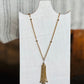 A gold-toned beaded chain necklace with a multi-strand tassel pendant, displayed on a white neck mannequin against a softly blurred indoor background.
