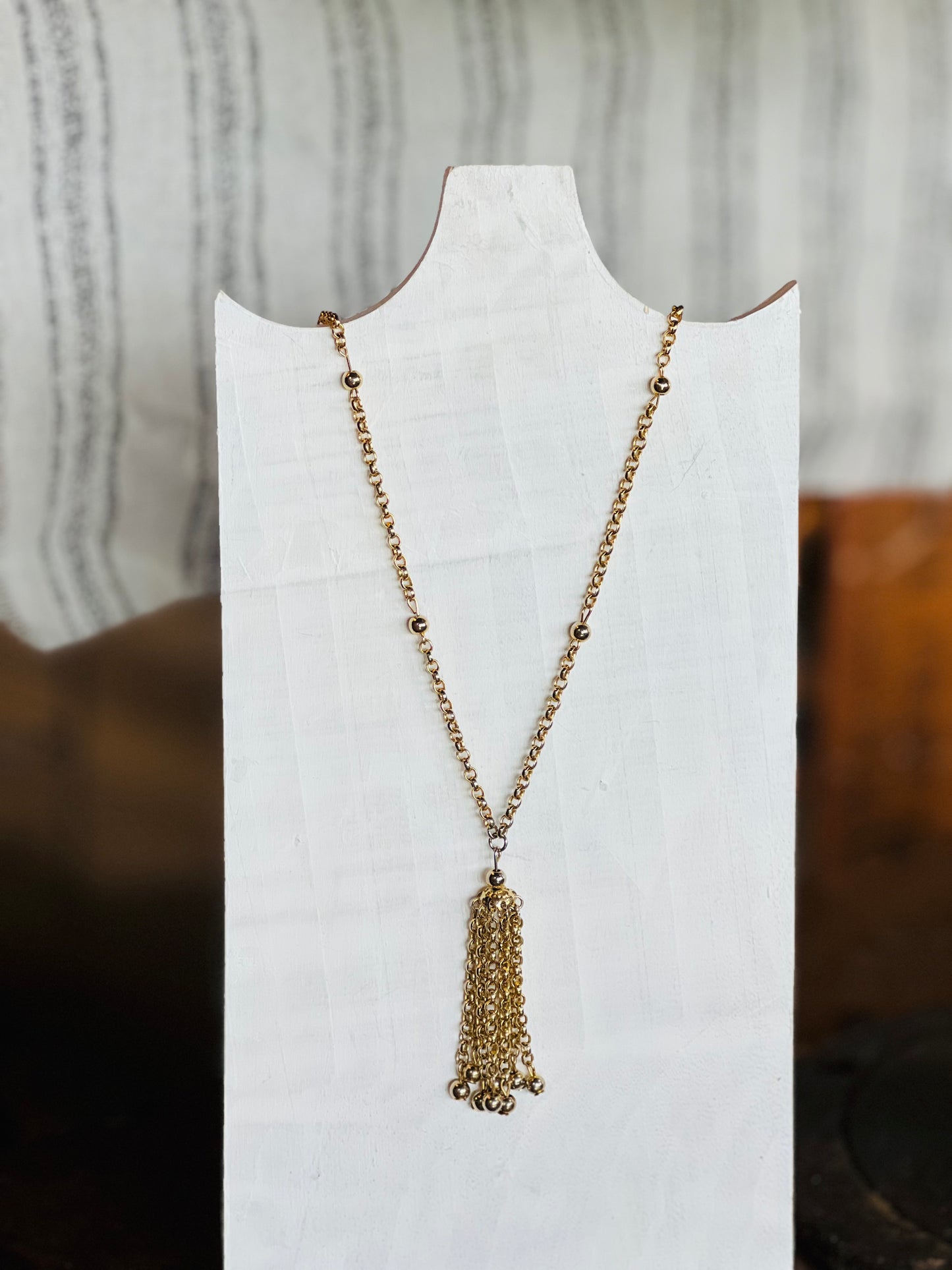 A gold-toned beaded chain necklace with a multi-strand tassel pendant, displayed on a white neck mannequin against a softly blurred indoor background.