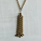 A gold-tone chain necklace with a long, multi-strand tassel pendant, displayed against a textured white background.