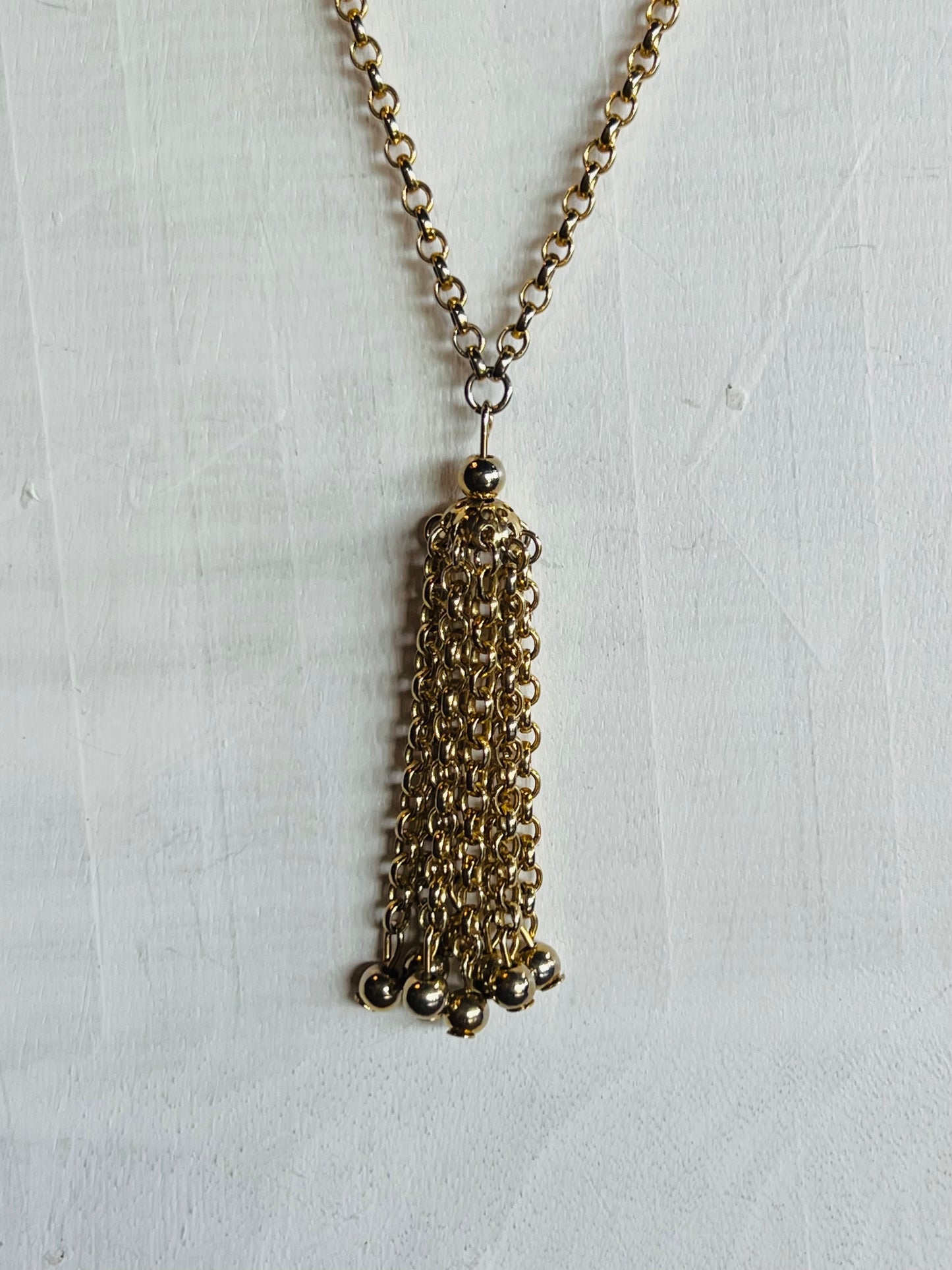 A gold-tone chain necklace with a long, multi-strand tassel pendant, displayed against a textured white background.