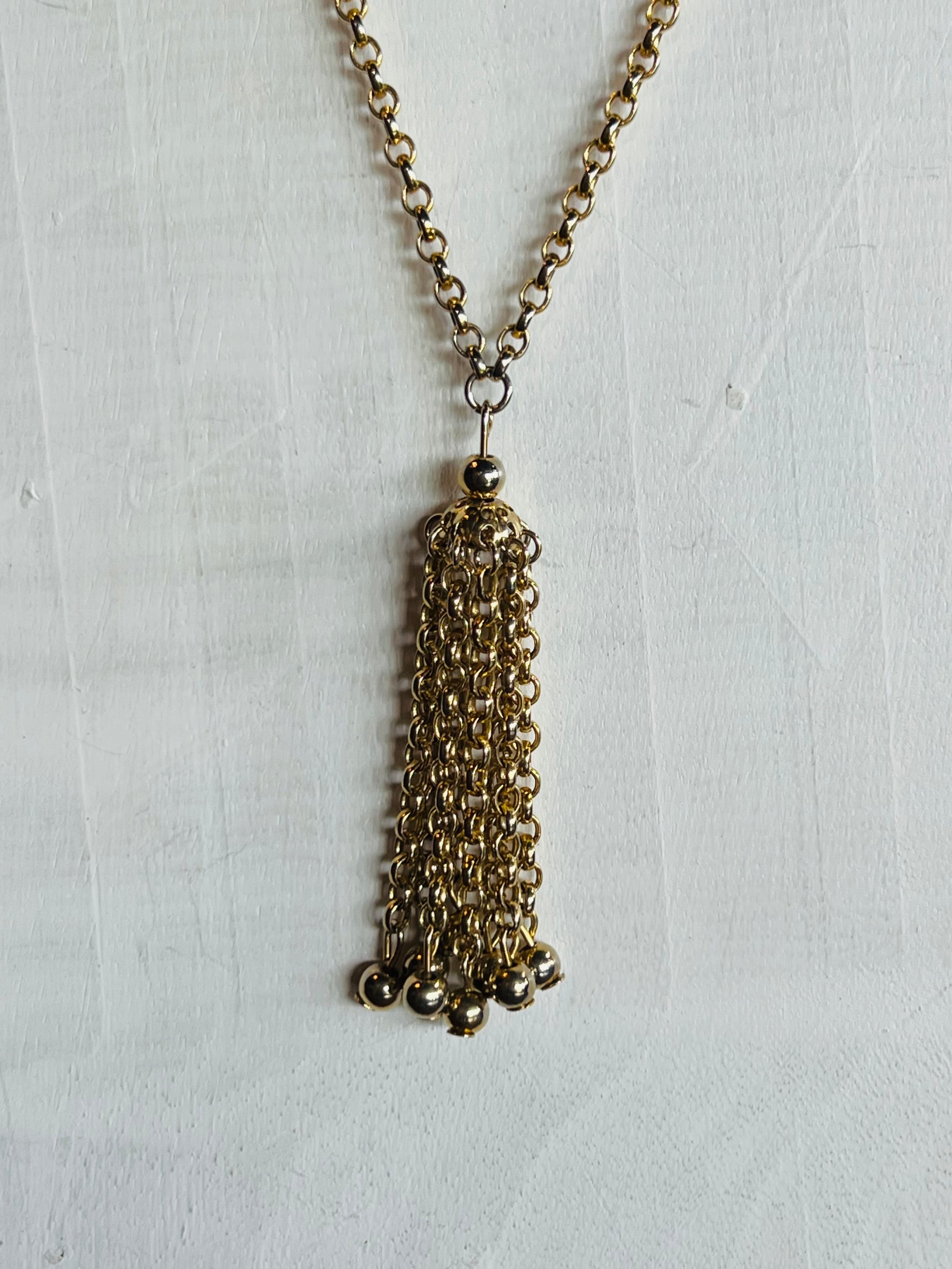 A gold-tone chain necklace with a long, multi-strand tassel pendant, displayed against a textured white background.