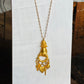 A gold pendant necklace featuring a stylized hand holding a ring, with small dangling charms including a heart and floral details, displayed on a white fabric-covered stand.