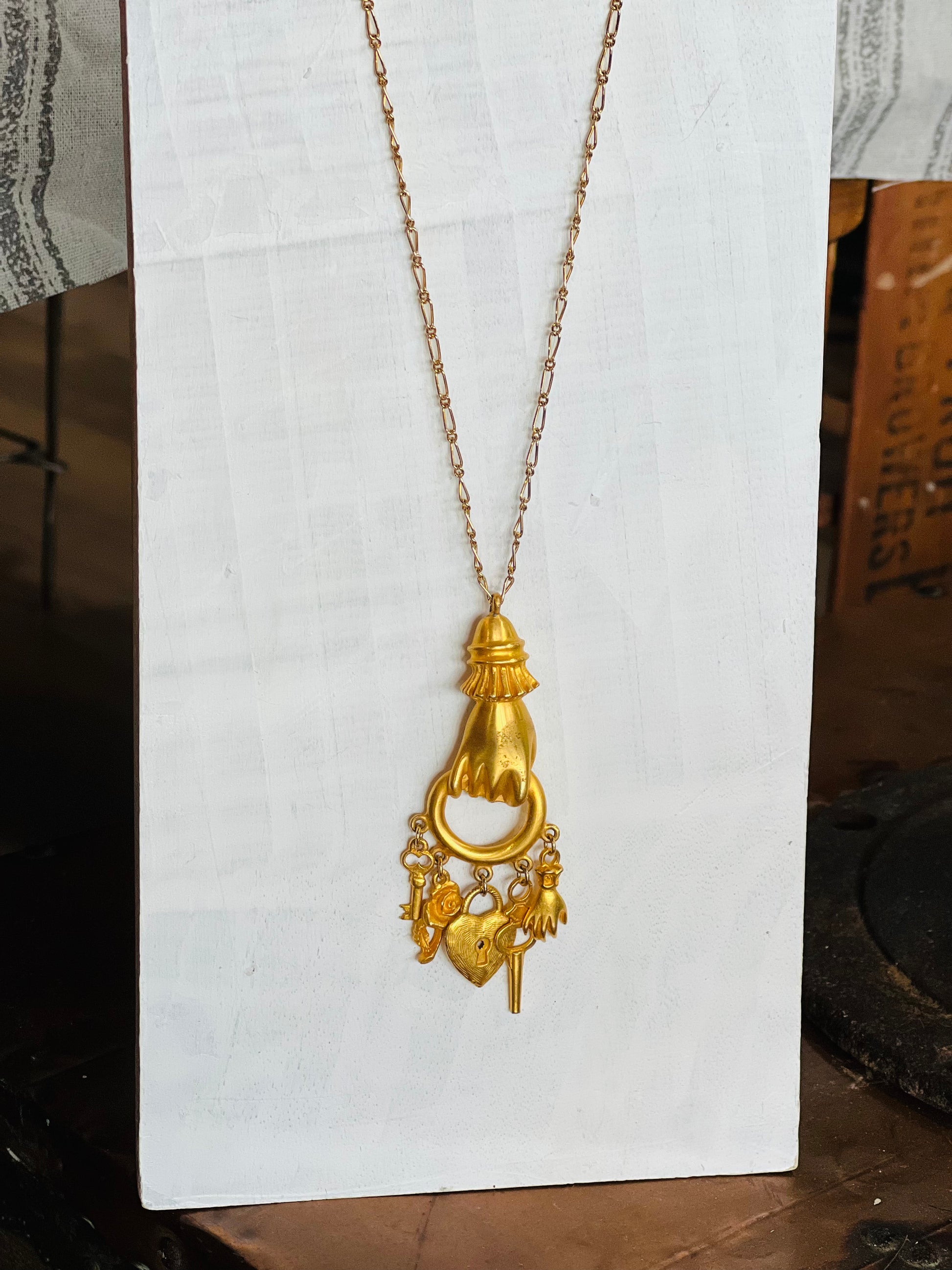 A gold pendant necklace featuring a stylized hand holding a ring, with small dangling charms including a heart and floral details, displayed on a white fabric-covered stand.
