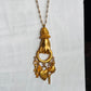 A gold-tone pendant necklace featuring a stylized hand holding a ring, with dangling charms including keys and a small heart, displayed against a white textured background.
