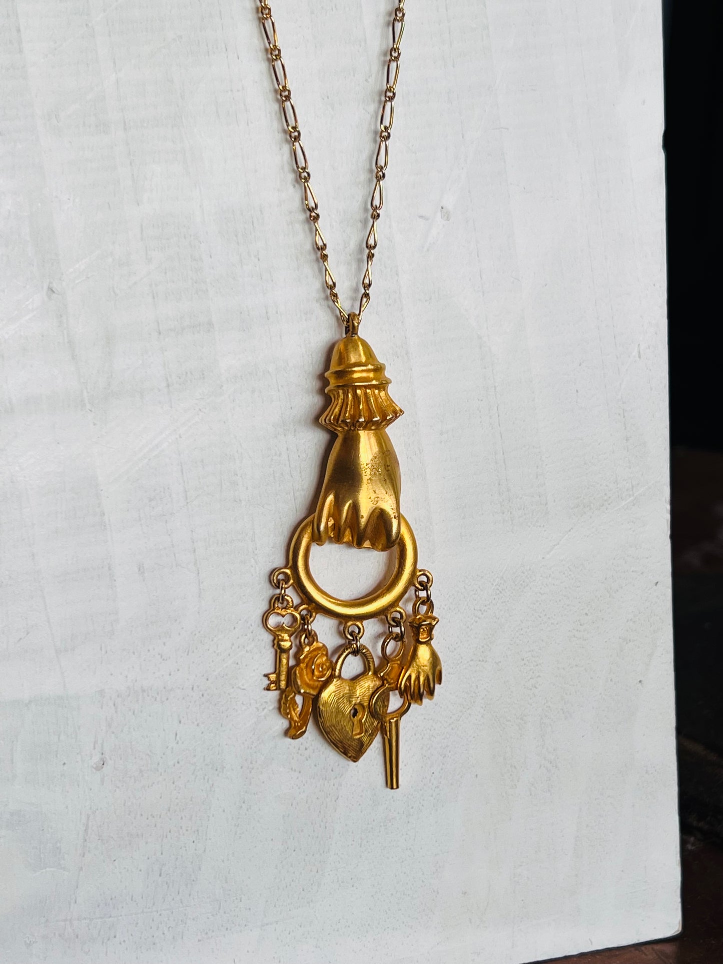 A gold-tone pendant necklace featuring a stylized hand holding a ring, with dangling charms including keys and a small heart, displayed against a white textured background.