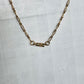 A gold-toned chain necklace with a central clasp, laid flat on a light gray textured surface.