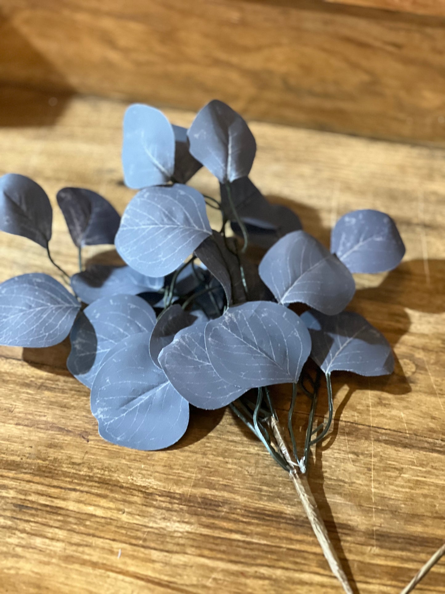 A cluster of artificial dusty blue-gray hydrangea-like leaves on a thin stem, displayed on a wooden tabletop.