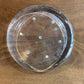 A clear circular plastic tray with eight evenly spaced small holes around the base, photographed from above on a brown wooden surface.