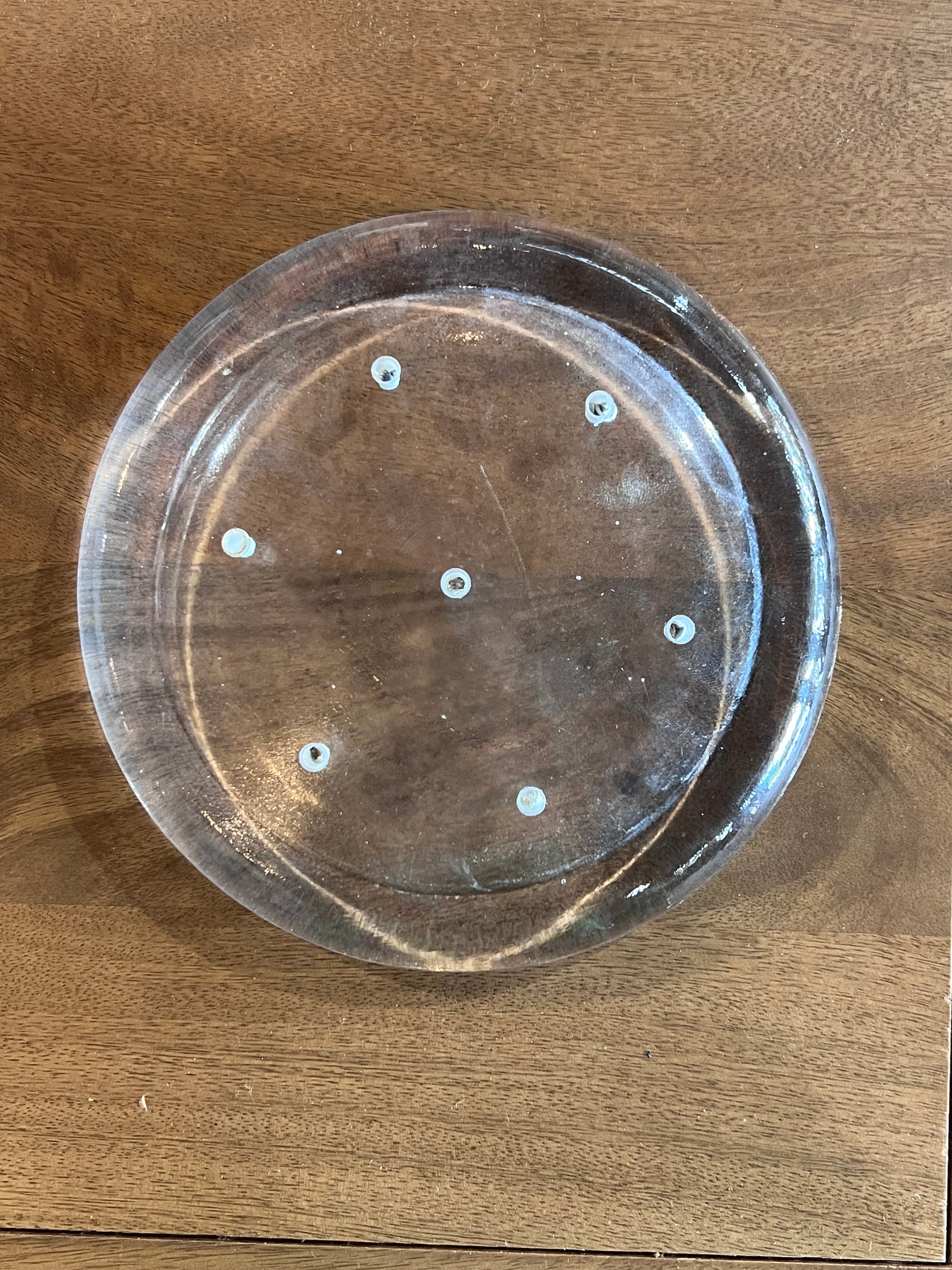 A clear circular plastic tray with eight evenly spaced small holes around the base, photographed from above on a brown wooden surface.