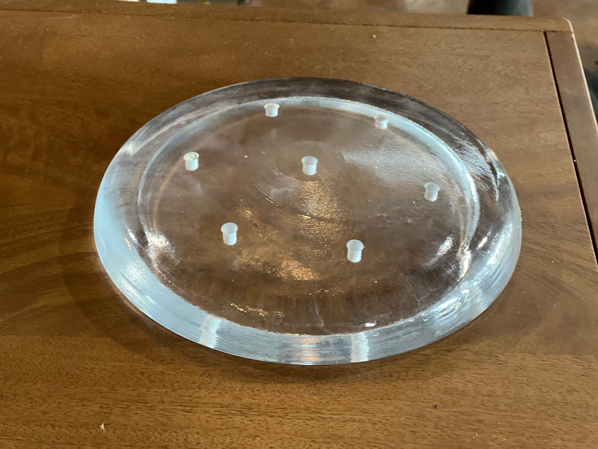 A clear, round acrylic or glass trivet with six small raised feet, shown on a wooden tabletop.