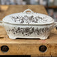 A white ceramic lidded casserole dish with a dark floral pattern, resting on a rustic wooden workbench.
