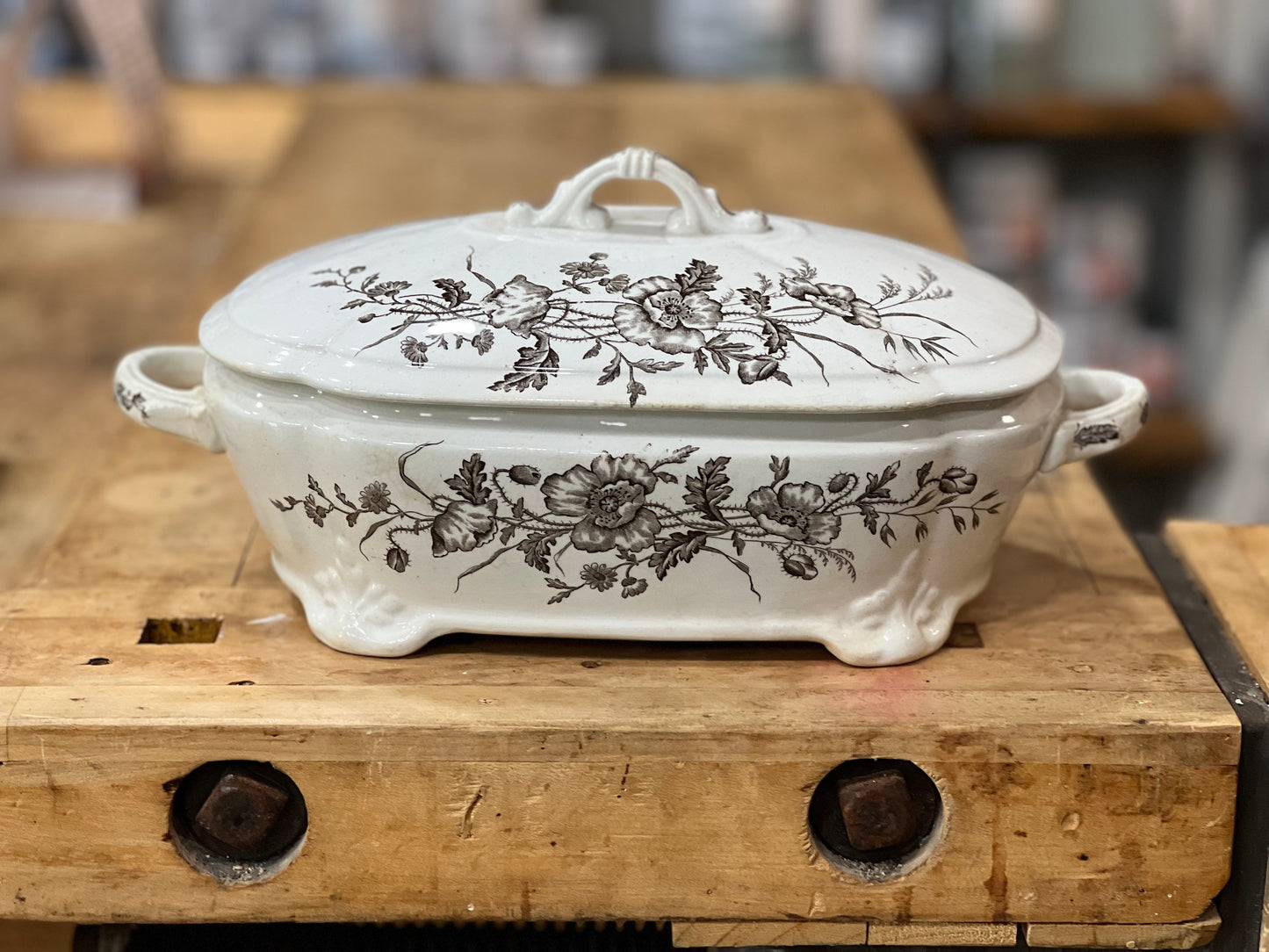 A white ceramic lidded casserole dish with a dark floral pattern, resting on a rustic wooden workbench.