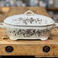 A white ceramic lidded casserole dish with a brown floral toile pattern, resting on a rustic wooden workbench.