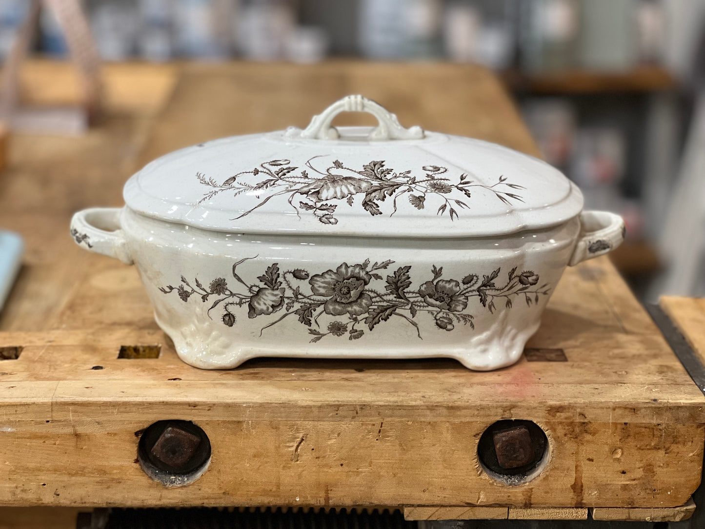 A white ceramic lidded casserole dish with a brown floral toile pattern, resting on a rustic wooden workbench.