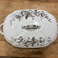 A white ceramic lidded casserole dish with a floral brown transfer pattern and side handles, photographed from above on a wooden tabletop.
