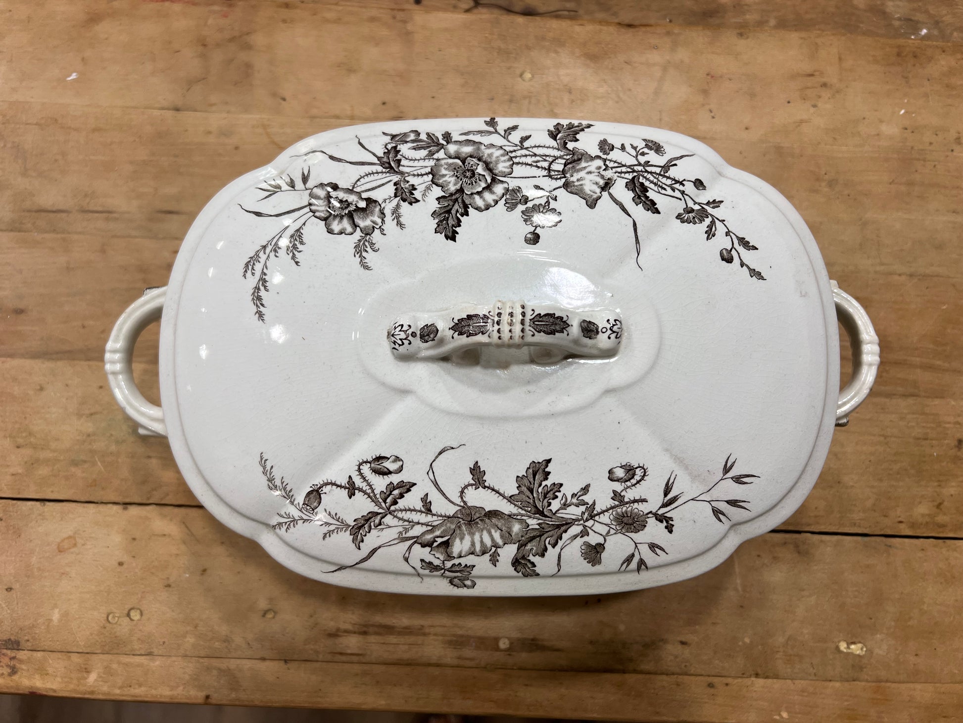 A white ceramic lidded casserole dish with a floral brown transfer pattern and side handles, photographed from above on a wooden tabletop.