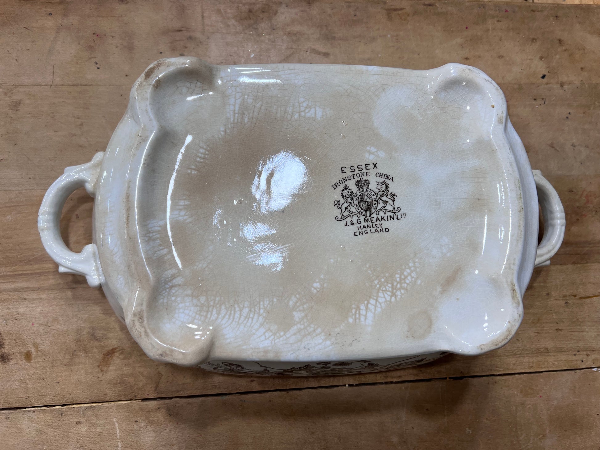 An antique ceramic baking dish shown from the bottom, featuring a glossy off-white glaze, two side handles, and a central black printed 'ESSEX' brand mark, placed on a rustic wooden surface.