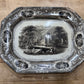 An antique octagonal ceramic serving tray with a black and white landscape illustration in the center and ornate decorative borders, displayed on a wooden surface.