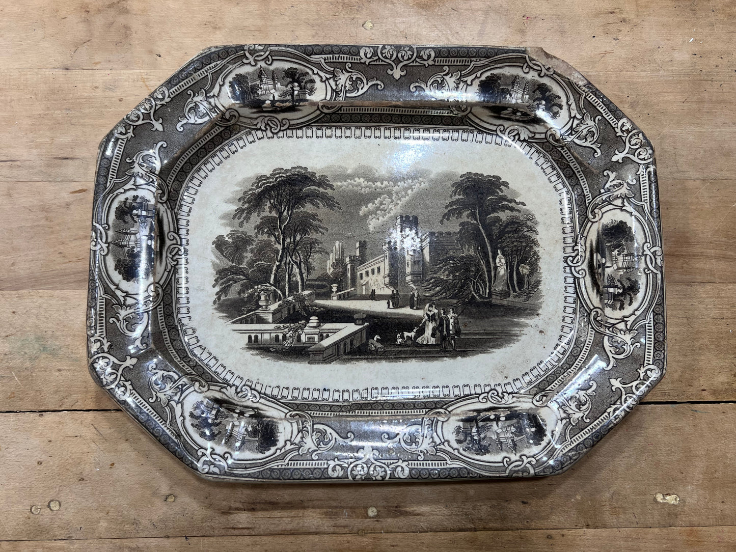 An antique octagonal ceramic serving tray with a black and white landscape illustration in the center and ornate decorative borders, displayed on a wooden surface.