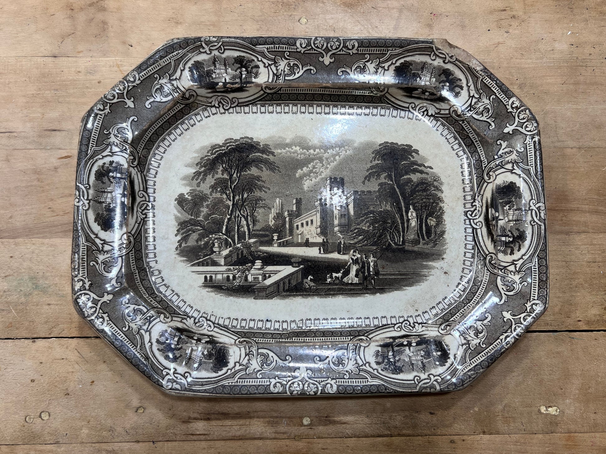 An antique octagonal ceramic serving tray with a black and white landscape illustration in the center and ornate decorative borders, displayed on a wooden surface.