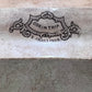 Close-up of a ceramic tile or stone surface with a decorative embossed or printed crest reading “CORINTHIA” and “E. CHALLINOR.