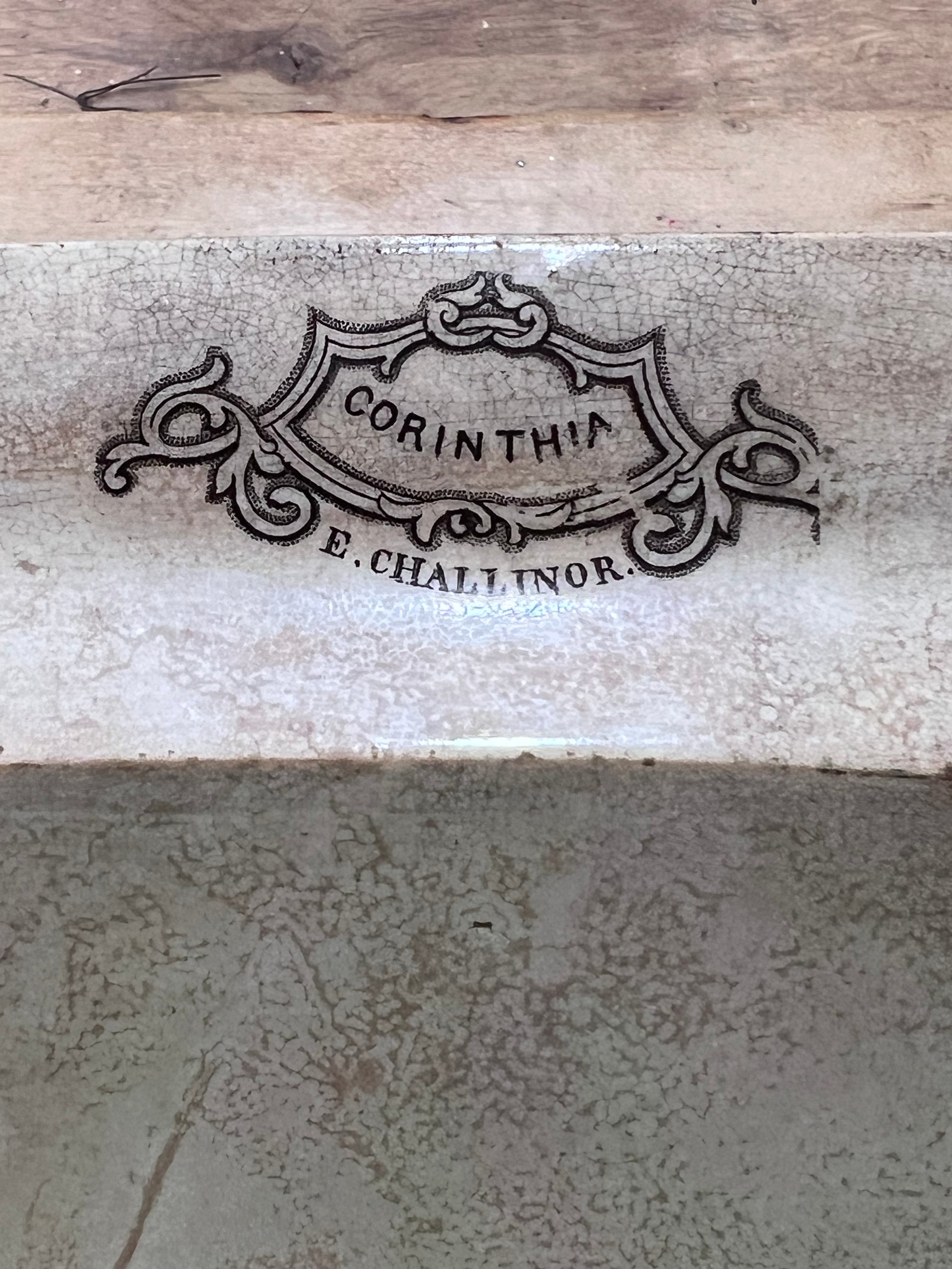 Close-up of a ceramic tile or stone surface with a decorative embossed or printed crest reading “CORINTHIA” and “E. CHALLINOR.