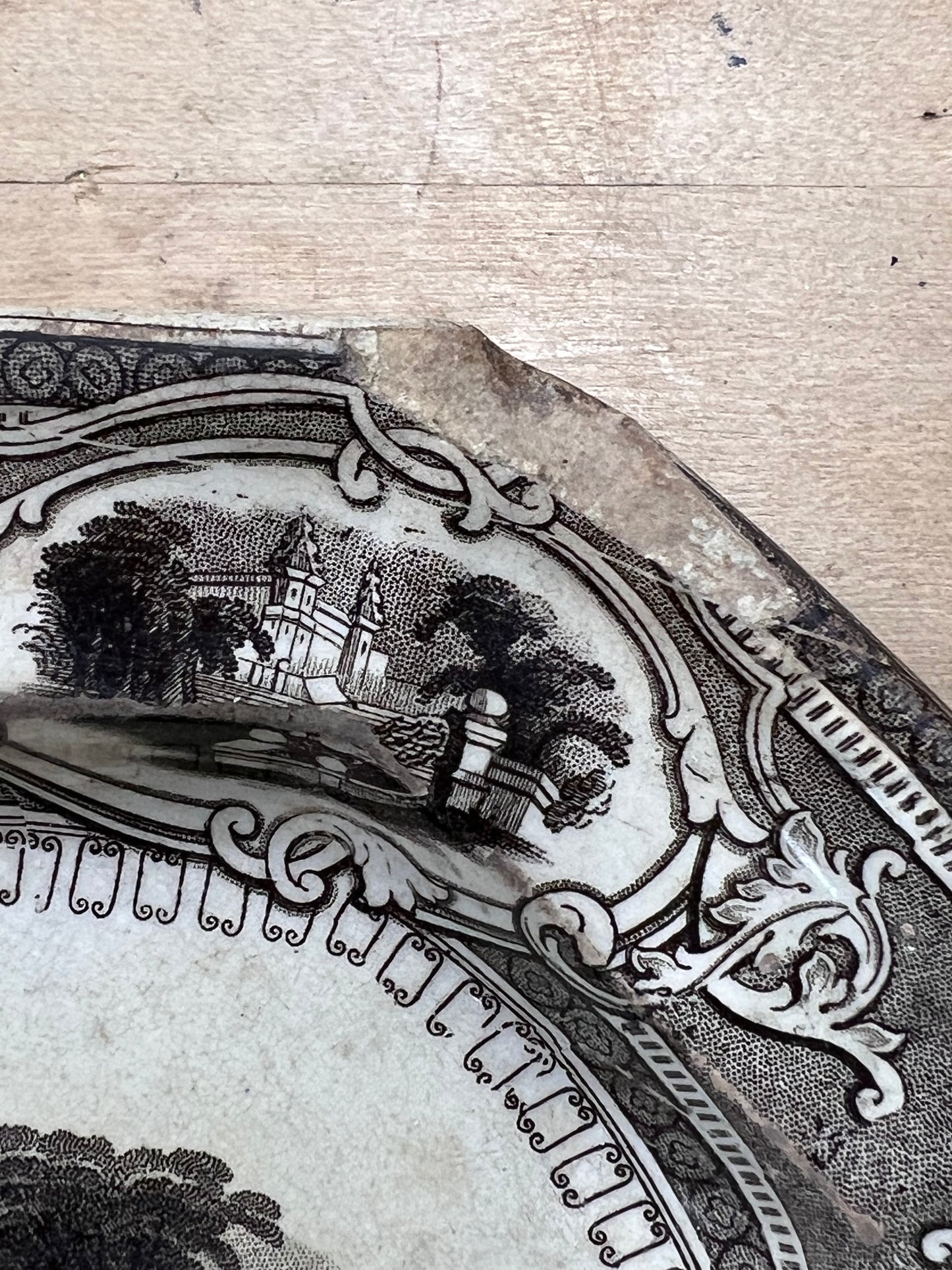 A close-up of a vintage ceramic plate or dish fragment with a black printed landscape scene and ornate border, resting on a light wood surface.