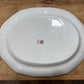 An oval white ceramic serving platter with a raised rim, photographed from above on a rustic wooden surface. A small brown brand mark is visible in the center.