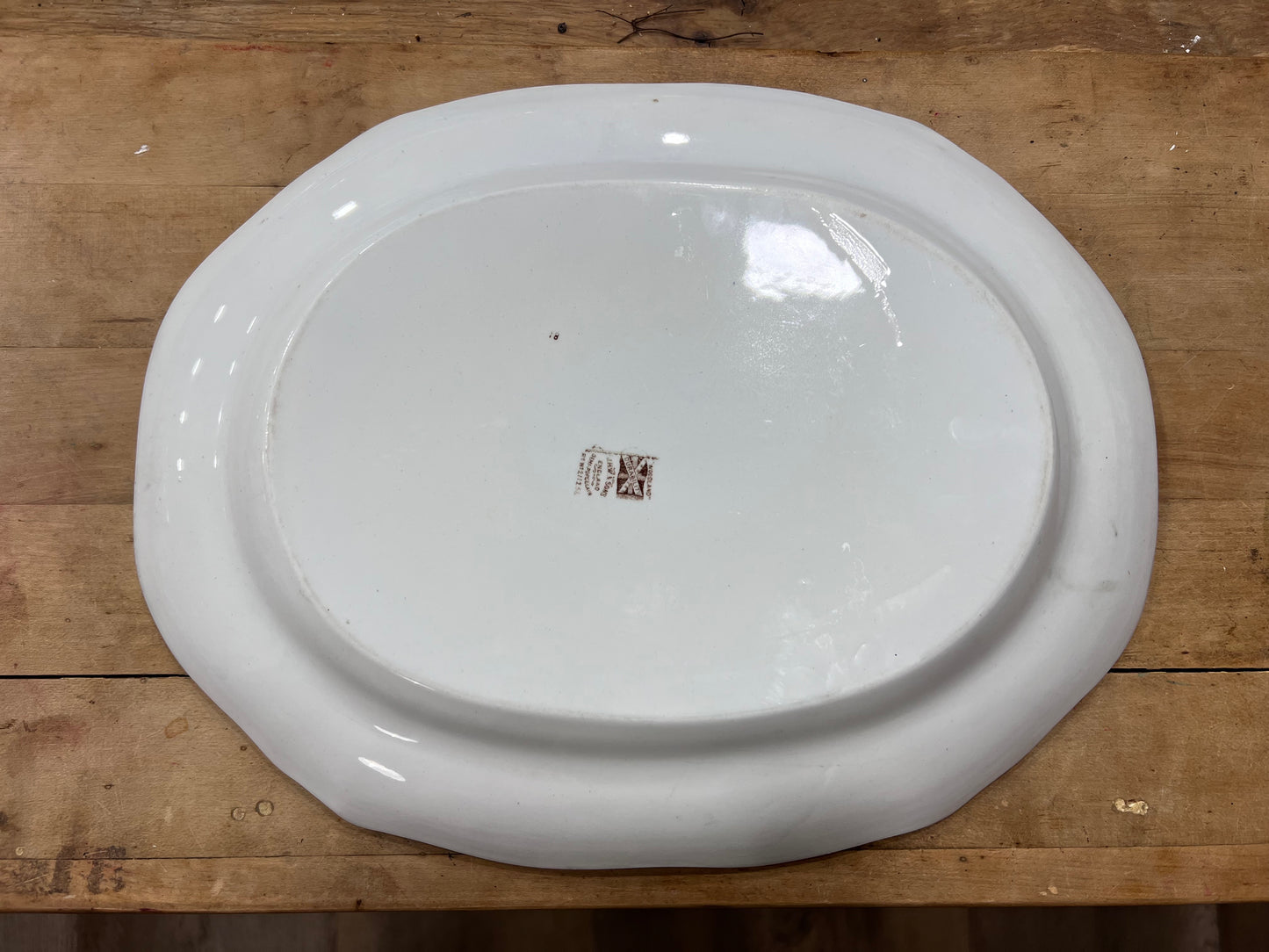 An oval white ceramic serving platter with a raised rim, photographed from above on a rustic wooden surface. A small brown brand mark is visible in the center.
