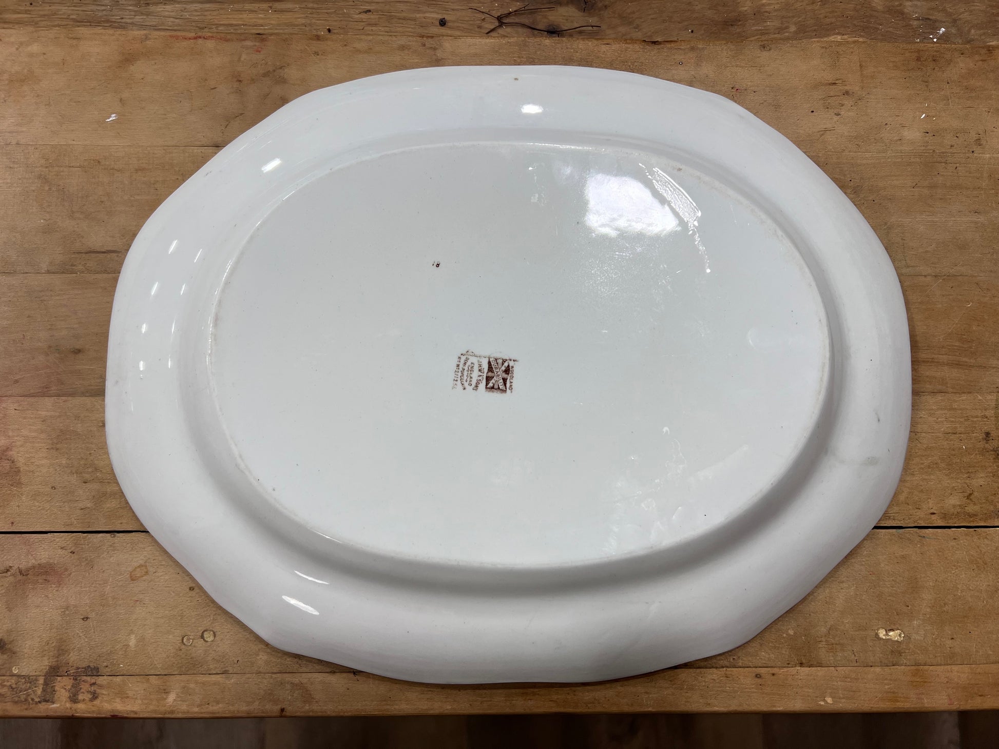 An oval white ceramic serving platter with a raised rim, photographed from above on a rustic wooden surface. A small brown brand mark is visible in the center.