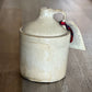 A handmade ceramic stoneware jug with a lid and handle, featuring a speckled cream glaze and a small red and black ribbon tag attached, set on a wooden floor.