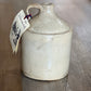 A vintage ceramic milk jug with a rounded body, small handle, and a hanging paper tag, set on a wooden surface.