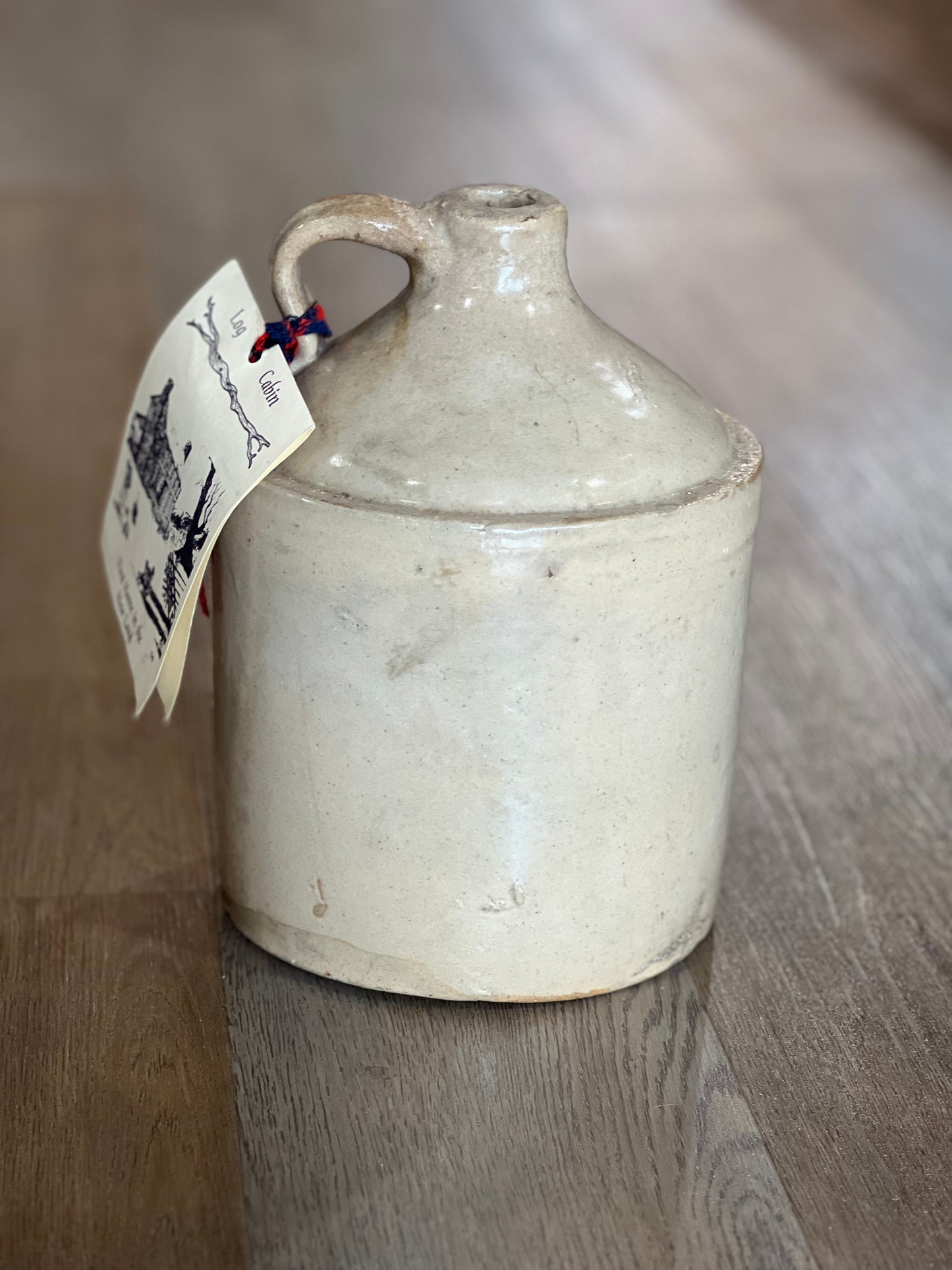 A vintage ceramic milk jug with a rounded body, small handle, and a hanging paper tag, set on a wooden surface.