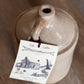 A rustic beige ceramic jug with a handle and spout, photographed on a wooden surface. A paper tag hangs from the handle with a red string, printed with the words 'Log Cabin' and a black line drawing of a cabin.