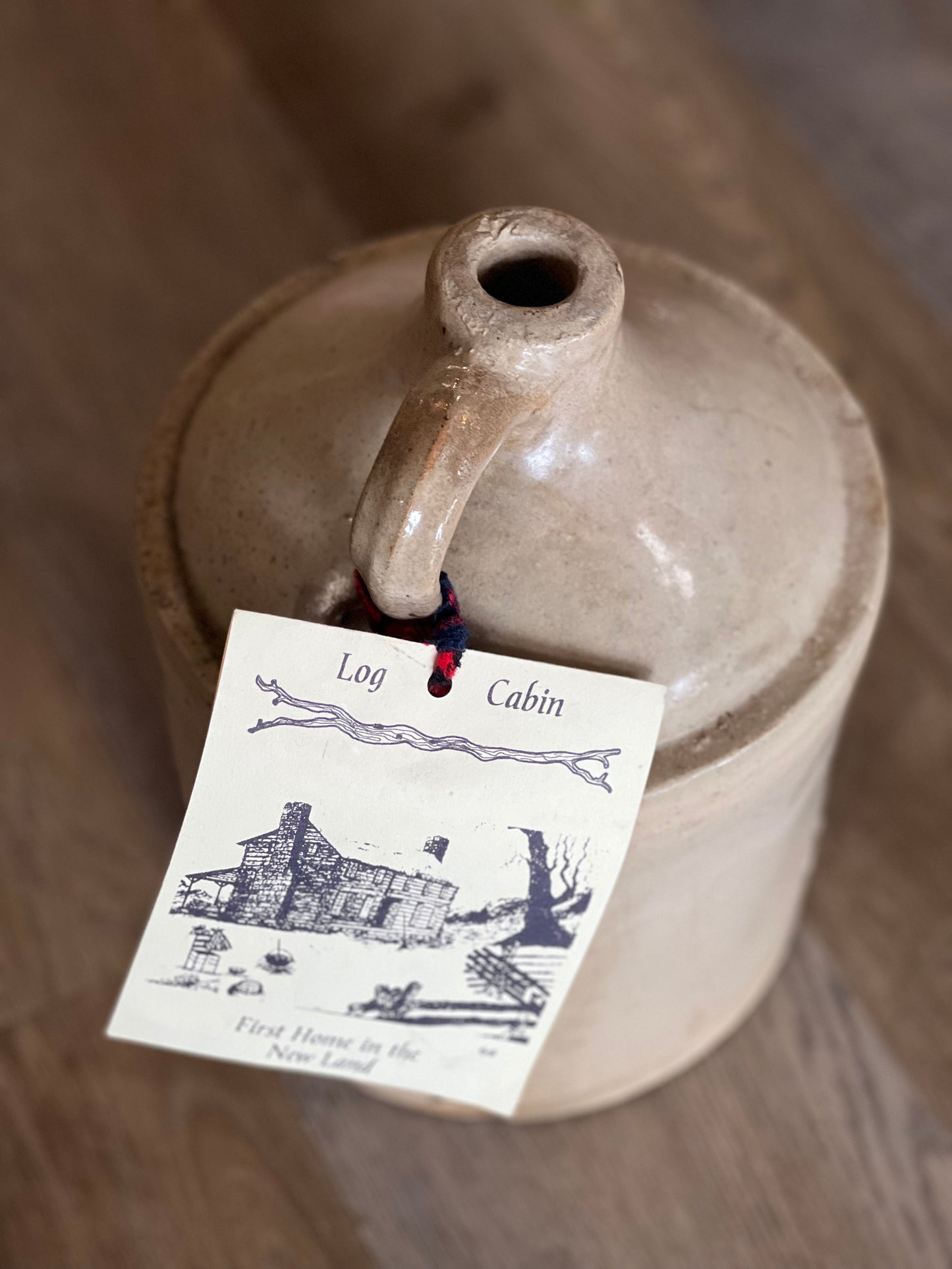 A rustic beige ceramic jug with a handle and spout, photographed on a wooden surface. A paper tag hangs from the handle with a red string, printed with the words 'Log Cabin' and a black line drawing of a cabin.