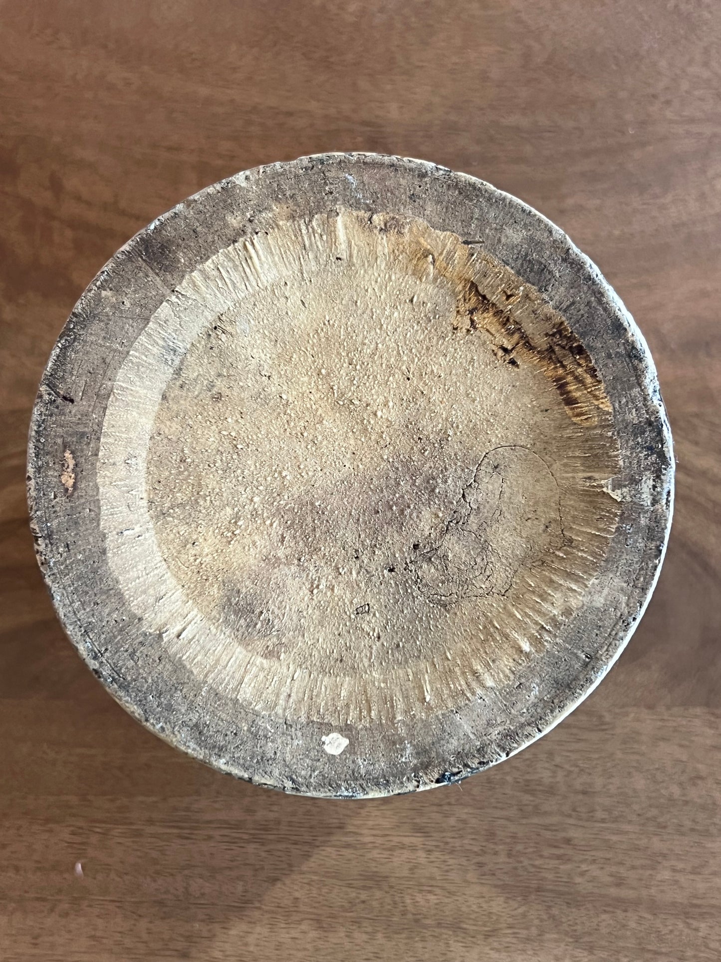 A round, rustic wooden bowl or dish with a weathered, textured surface, photographed from directly above on a brown wooden tabletop.