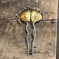 A pair of vintage-style hairpins with gold-toned leaf-shaped tops, small clear rhinestone accents, and wavy silver-toned stems, displayed on a rustic wooden surface.