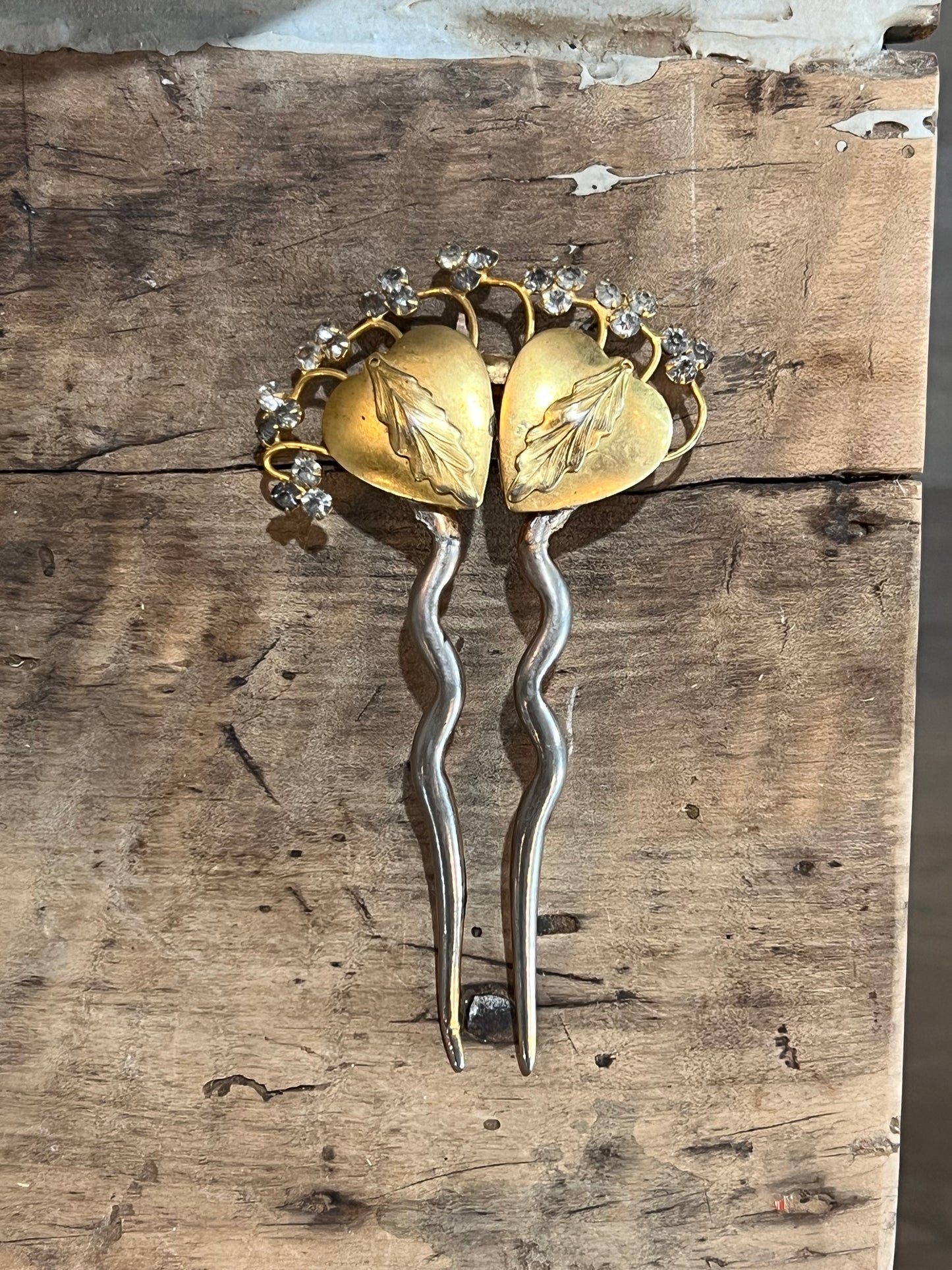 A pair of vintage-style hairpins with gold-toned leaf-shaped tops, small clear rhinestone accents, and wavy silver-toned stems, displayed on a rustic wooden surface.