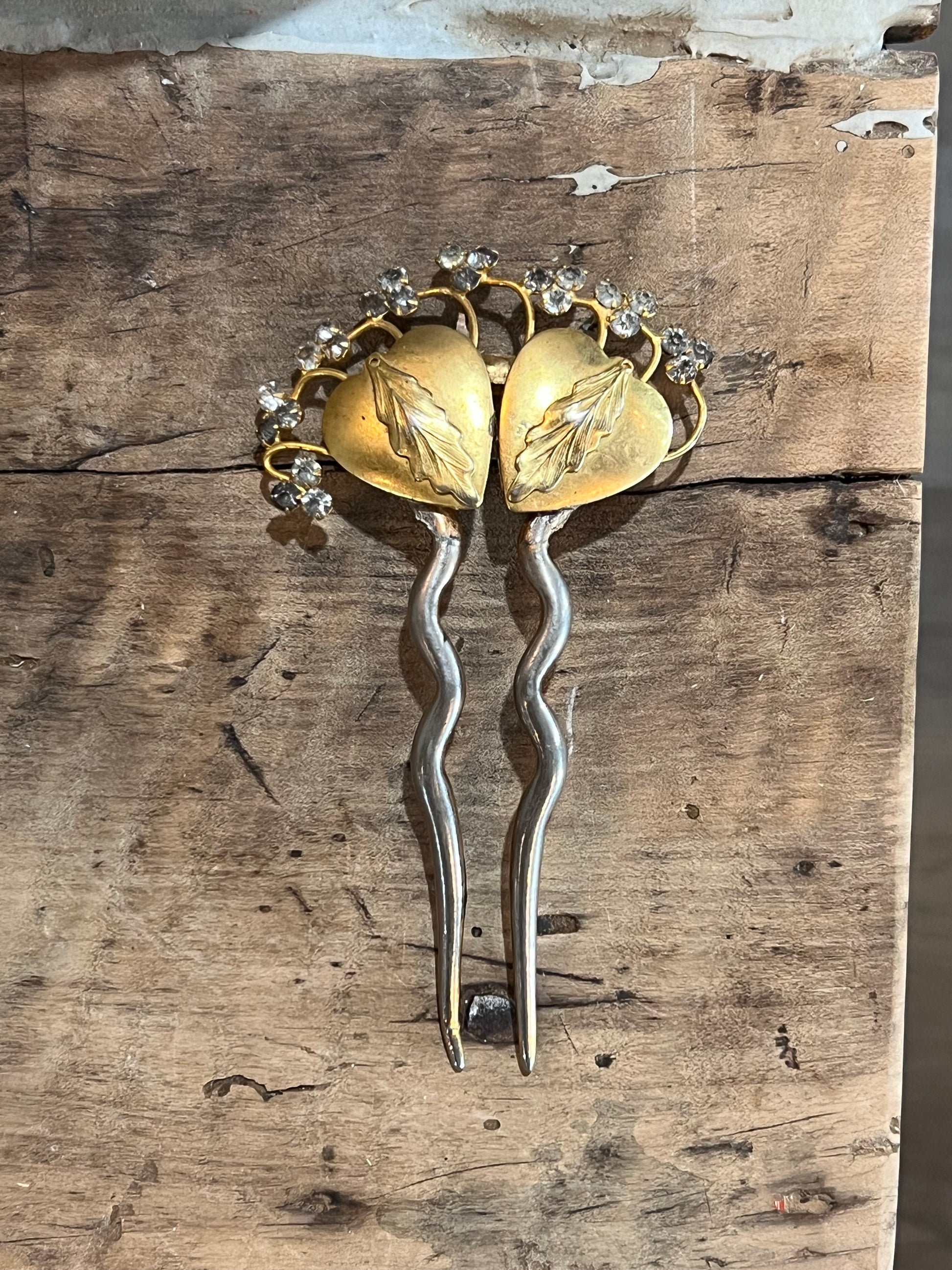 A pair of vintage-style hairpins with gold-toned leaf-shaped tops, small clear rhinestone accents, and wavy silver-toned stems, displayed on a rustic wooden surface.