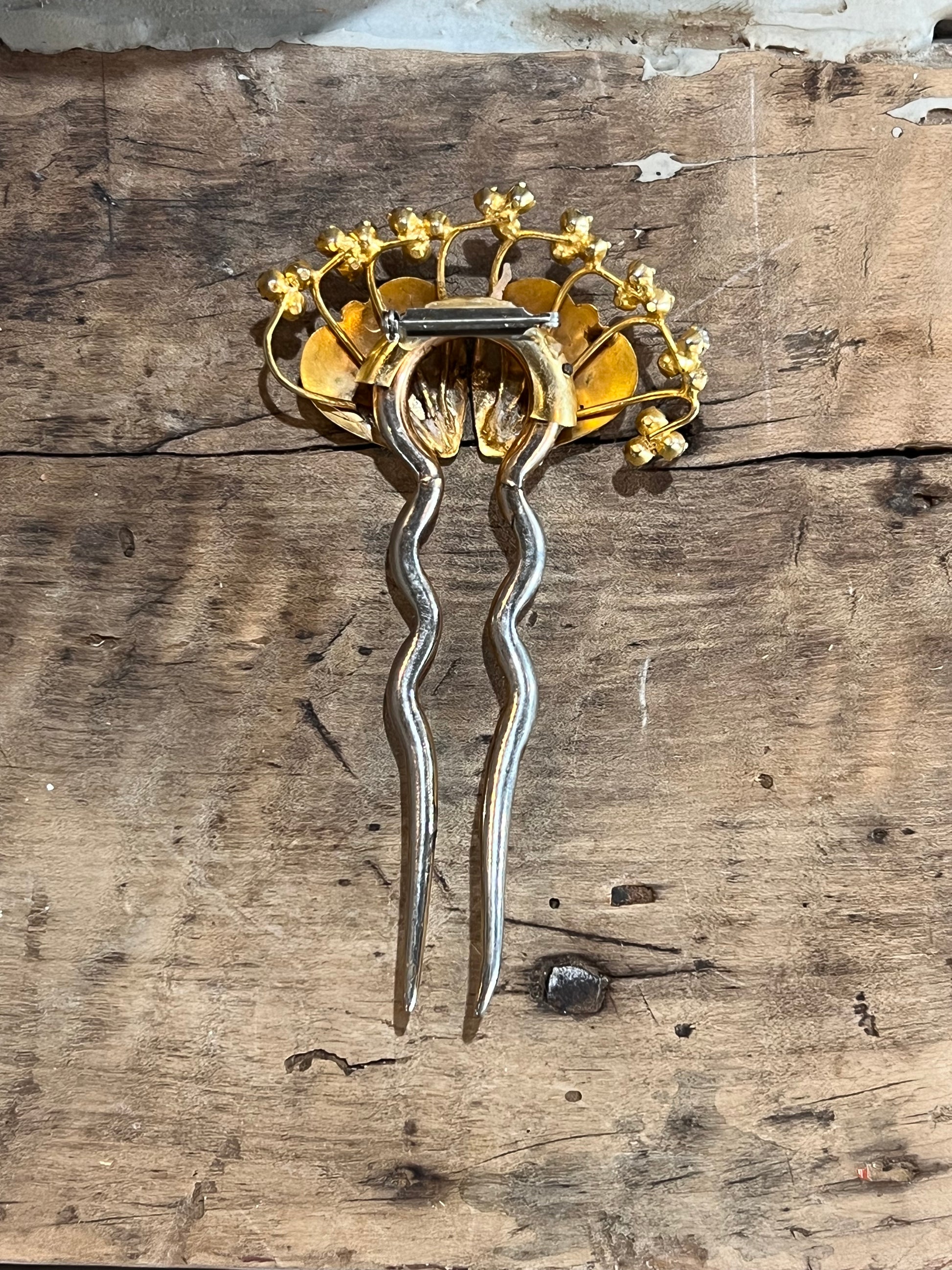 A vintage gold and silver filigree hair pin with a crown-like top and two long twisted stems, displayed on a rustic wooden surface.