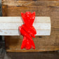 A glossy bright red sculptural ring with a twisted, ribbon-like form, displayed on a light-colored wooden dowel against a rustic wooden background.