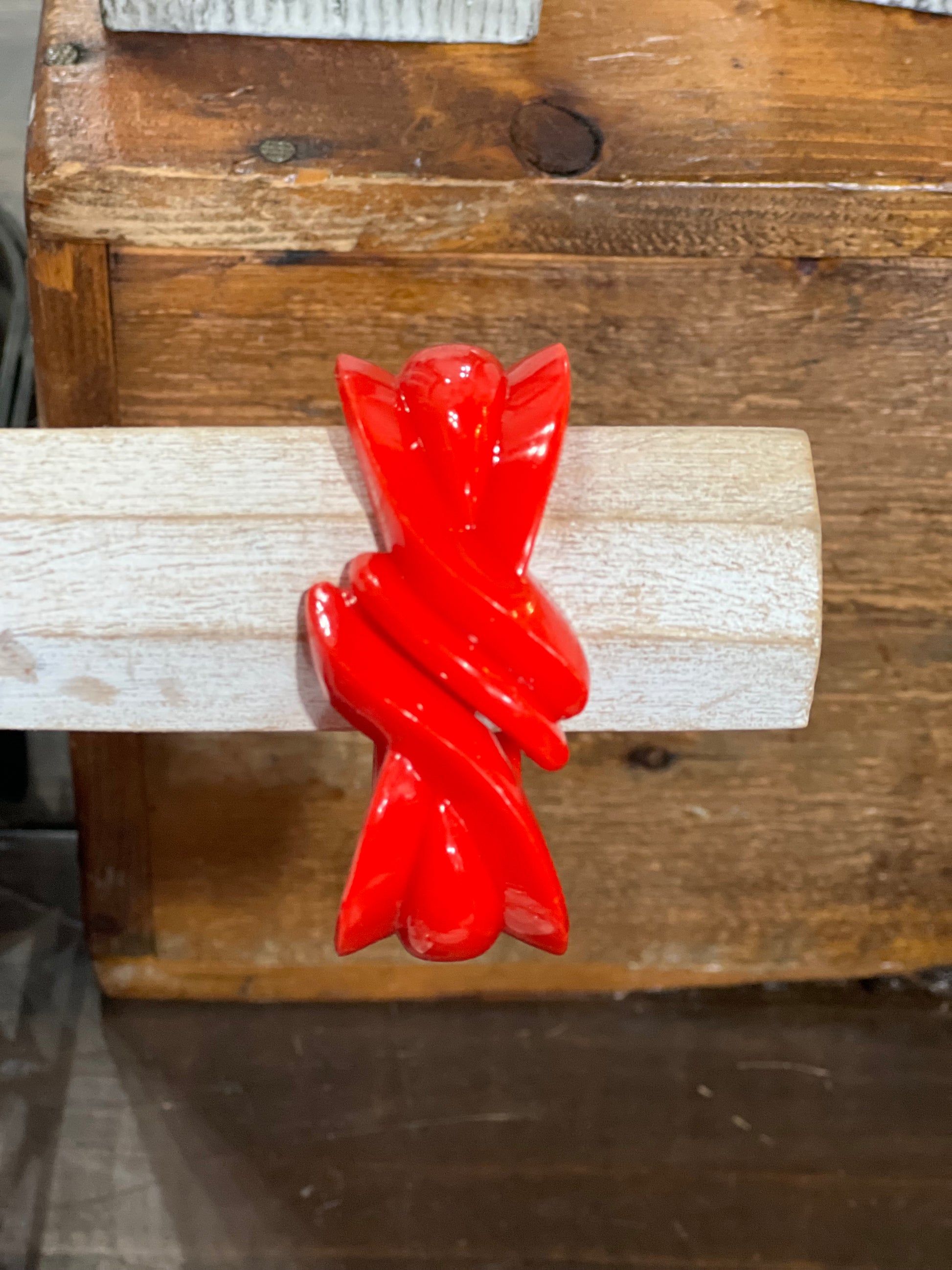 A glossy bright red sculptural ring with a twisted, ribbon-like form, displayed on a light-colored wooden dowel against a rustic wooden background.