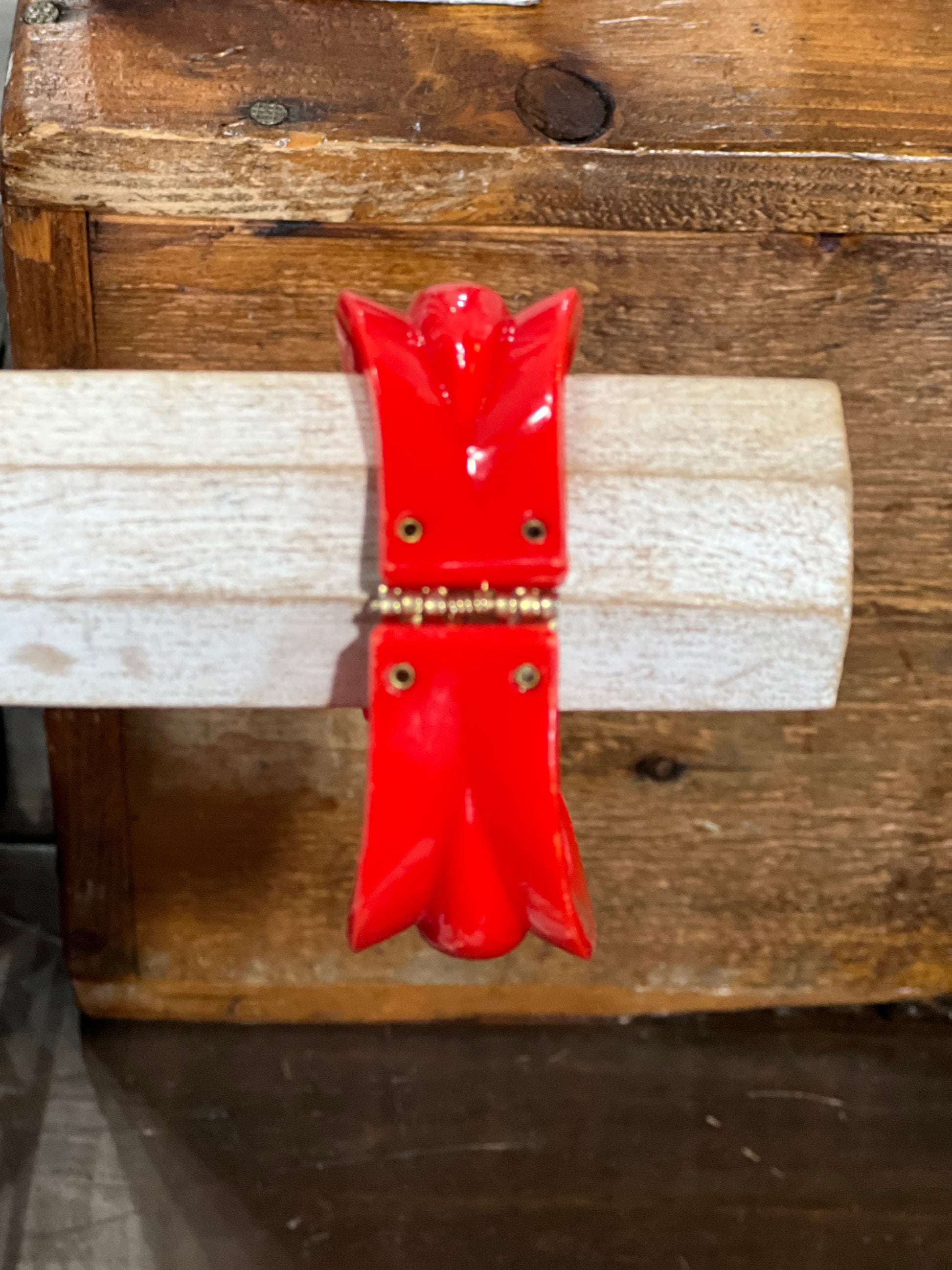 A glossy red decorative wall hook or hanging ornament with a small gold-toned metal accent, shown mounted on a light wood dowel against a rustic wooden background.